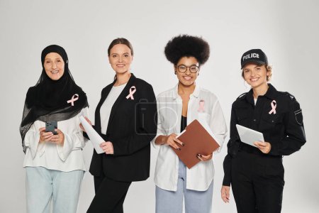 lächelnd verschiedene interrassische Frauen mit rosa Schleifen, die Geräte und Papiere isoliert auf grau halten