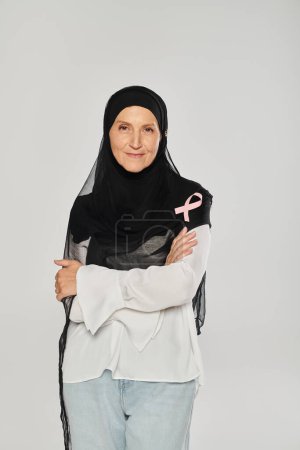 smiling woman in hijab with pink ribbon of breast cancer awareness isolated on grey