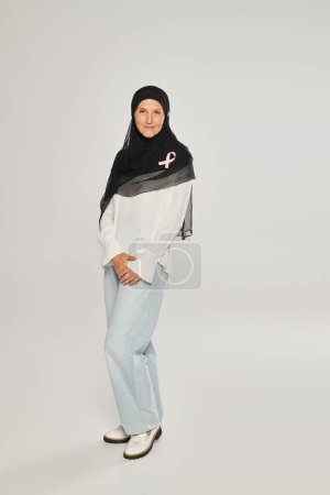 Photo for Smiling woman in hijab with pink ribbon of breast cancer awareness standing on grey background - Royalty Free Image