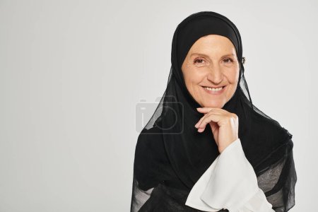 Porträt einer fröhlichen Frau im Hijab, die in die Kamera blickt, während sie isoliert auf grau posiert