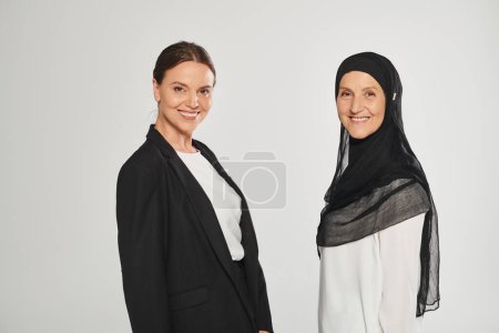 smiling businesswoman and woman in hijab looking at camera together isolated on grey