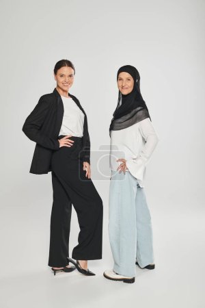 longitud completa de la sonriente mujer de negocios y mujer en hijab posando sobre fondo gris