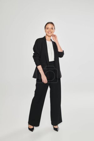 full length of cheerful woman in heels and suit standing on grey background