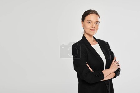 selbstbewusste brünette Frau im Anzug verschränkt die Arme und blickt isoliert in die Kamera auf grau