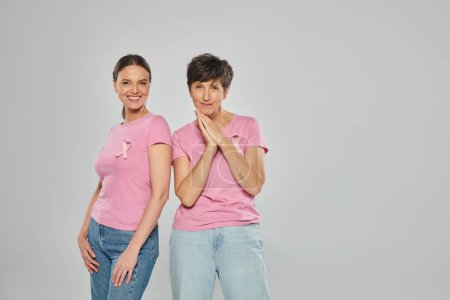 smiling women with pink ribbons and t-shirts standing isolated on grey, breast cancer