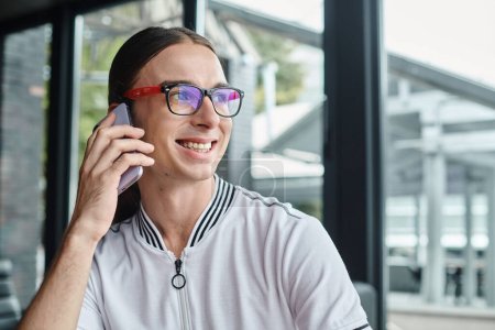 close up cheerful young man in glasses talking on phone with glass background, coworking concept mug #673733026