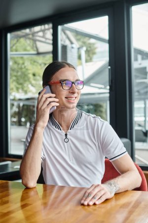 young man with pony tail and glasses talking on phone at table with glass on backdrop, coworking