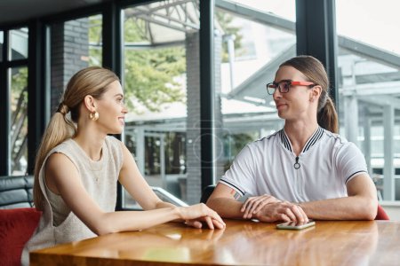 junge Mitarbeiter Mann und Frau plaudern und schauen sich vor Glaskulisse an, Coworking