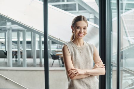 fröhliche junge Frau in Business-Freizeitkleidung blickt in die Kamera mit Fensterkulisse, Coworking