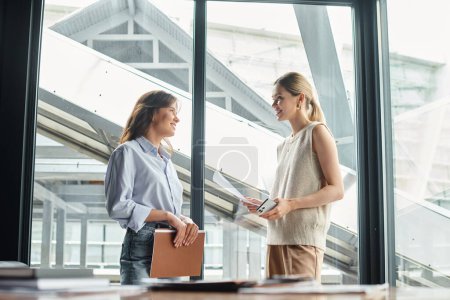 two female coworkers smiling and looking at each other, holding phone and working papers, coworking