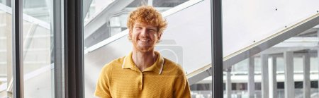 cheerful ginger man smiling sincerely looking at camera with window on background, coworking, banner