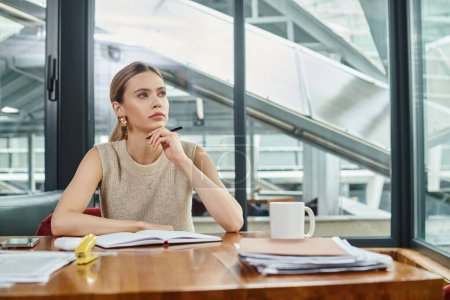 thoughtful woman in smart wear sitting at table working on documents, hand to chin, coworking