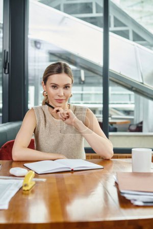 young attractive woman with ponytail working on her papers and looking at camera, coworking concept
