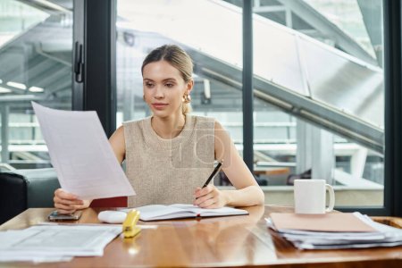 Téléchargez les photos : Jeune employée en tenue décontractée d'affaires assise à table et regardant sa paperasse, coworking - en image libre de droit