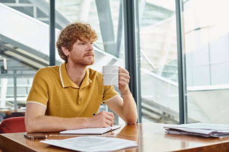 Nachdenklicher junger Mann mit roten Haaren, der an seinen Dokumenten arbeitet und Teetasse in der Hand hält, Coworking-Konzept
