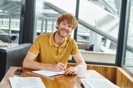 fröhlicher rothaariger Mann, der lächelt und in die Kamera schaut, während er an seinen Papieren arbeitet, Coworking-Konzept