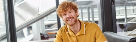 happy red haired man smiling and looking at camera with window backdrop, coworking concept, banner
