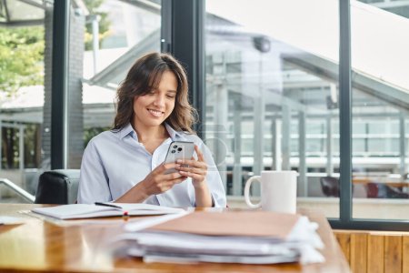 glückliche brünette Frau in smarter Kleidung, die am Tisch sitzt und auf das Smartphone schaut, Coworking-Konzept