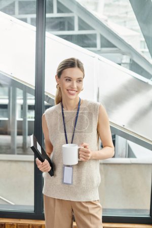 junge blonde Frau in schicker Kleidung mit Teetasse und Notizbuch, lächelnd und wegschauend, Coworking