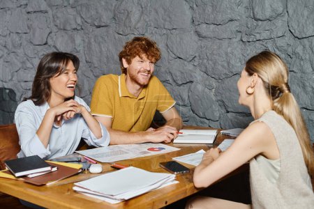 three cheerful coworkers laughing and looking at each other while working on papers, coworking