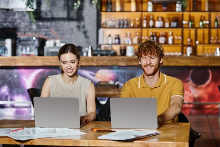 zwei gut gelaunte Kollegen lächeln, während sie an Laptops mit verschwommenem Balken vor dem Hintergrund arbeiten, Coworking
