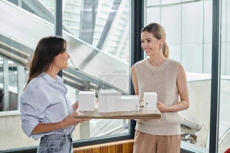 Team aus zwei jungen Frauen, die ein maßstabsgetreues Modell des Gebäudes halten und einander anschauen, Designbüro