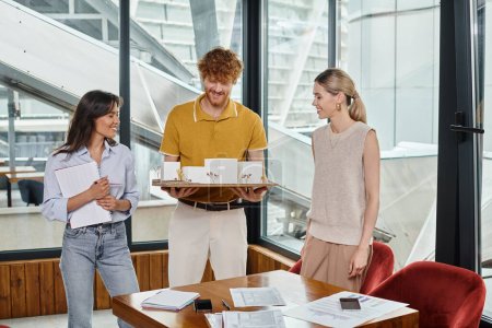 drei fröhliche junge Teammitglieder lächeln und betrachten das maßstabsgetreue Modell des Gebäudes, Designbüro