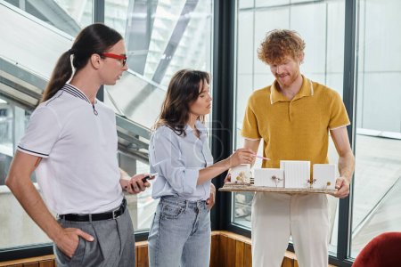 hochkonzentrierte Kollegen in smarter Freizeitkleidung, die an einem maßstabsgetreuen Modell des Gebäudes arbeiten, Designbüro
