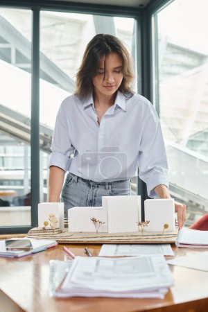 young brunette woman working on her documents and scale model with glass on backdrop, design bureau