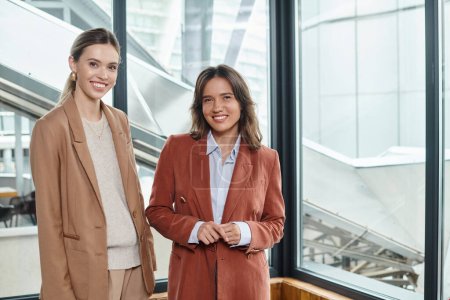 zwei Kolleginnen in eleganter, formaler Kleidung posieren auf Fensterhintergrund, Coworking-Konzept