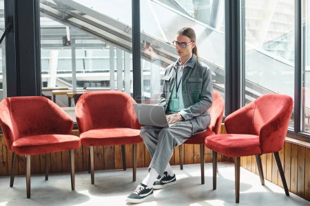 Foto de Joven en ropa inteligente se centró en su trabajo sentado en la silla con telón de fondo de vidrio, concepto de coworking - Imagen libre de derechos