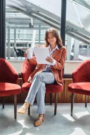 lächelnde junge Frau im braunen Blazer, die beim Tee ihre Unterlagen überprüft, Coworking