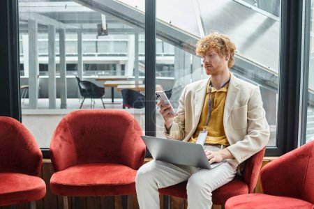 focused red haired man with laptop on his laps sitting and looking at his phone, coworking concept