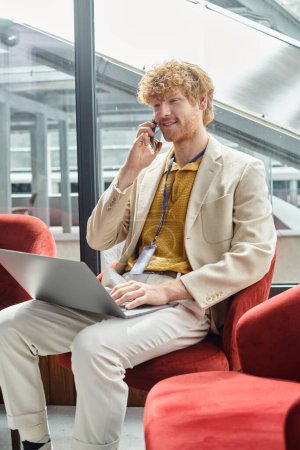 red haired jolly man holding his laptop and talking on mobile phone with glass backdrop, coworking