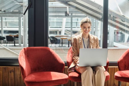 junge blonde Angestellte sitzt auf Stuhl und arbeitet an ihrem Laptop mit Glashintergrund, Coworking-Konzept