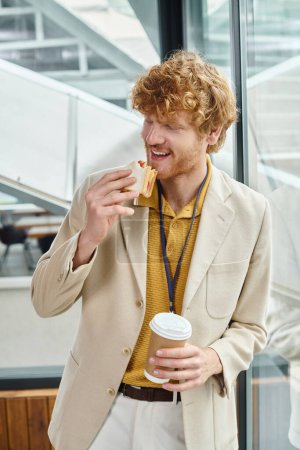 red haired man in smart attire enjoying his coffee and eating sandwich at lunch break, coworking