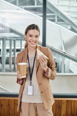 junge Frau in schicker Freizeitkleidung genießt Sandwich und Kaffee vor laufender Kamera, Coworking-Konzept