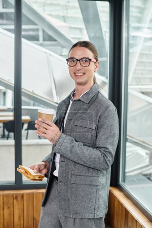 young man having lunch break eating sandwich drinking coffee with glass on backdrop, coworking