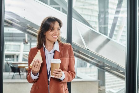 alegre joven mujer en ropa inteligente disfrutando de sándwich y café y mirando hacia otro lado, concepto de coworking