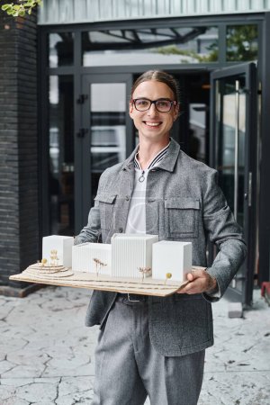 joven hombre alegre en gafas con modelo a escala de edificio mirando a la cámara, oficina de diseño