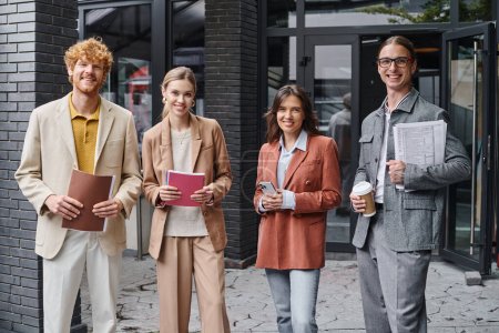 equipo creativo joven posando afuera sonriendo ante la cámara sosteniendo papeles, teléfono y taza de café, coworking