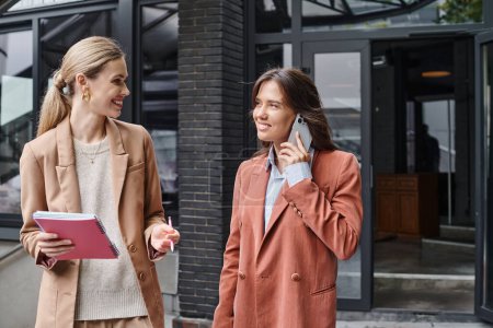 young female coworkers standing outside holding papers and talking on phone, coworking concept