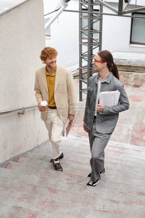 zwei junge männliche Kollegen gehen die Treppe hinauf, reden und lächeln sich an, Coworking-Konzept