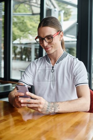 Nahaufnahme eines fröhlichen jungen Mannes mit Brille, der auf ein Handy mit Glashintergrund blickt, Coworking-Konzept