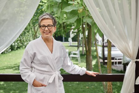 happy mature woman in glasses and white robe standing with hand in pocket during wellness retreat