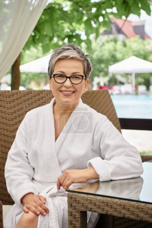 happy middle aged woman in glasses and white robe sitting at rattan table during wellness retreat
