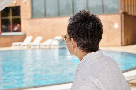 Reife Frau in Sonnenbrille und Bademantel ruht im Urlaub in privatem Pavillon neben Swimmingpool