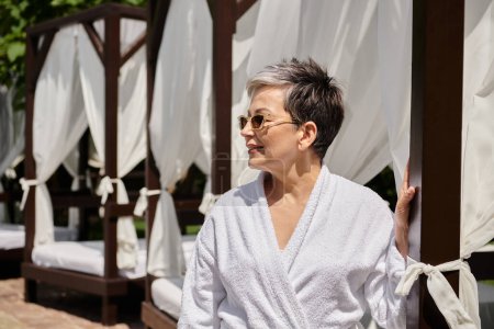 happy mature woman in sunglasses and robe resting in private pavilion during wellness retreat