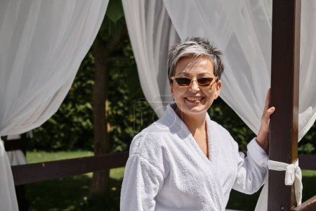 freudige reife Frau mit Sonnenbrille, die sich im privaten Pavillon ausruht, Wellness-Rückzugsort im Luxus-Resort