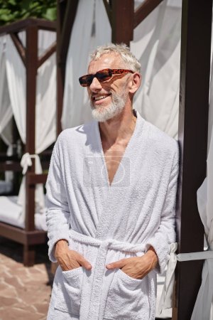 fröhlicher älterer Mann in Sonnenbrille und Robe posiert mit den Händen in den Taschen in der Nähe des privaten Pavillons